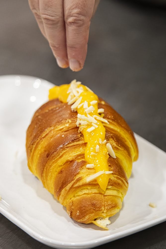 Croissants com recheio de ovos moles, com uma mão a colocar delicadamente amêndoas laminadas em cima do produto
